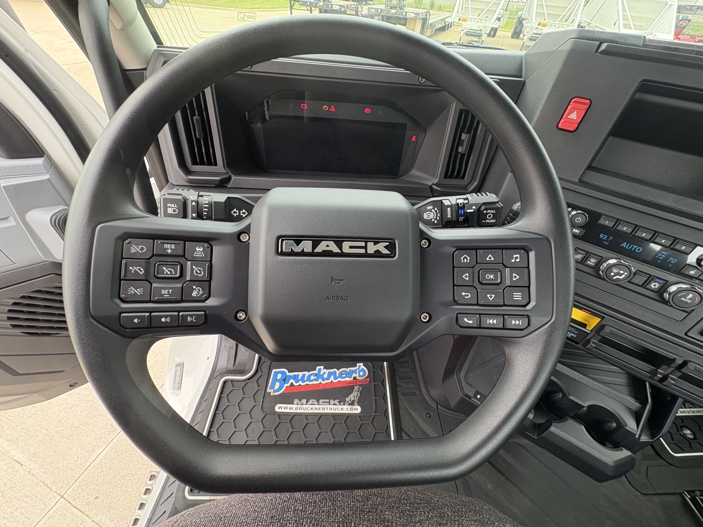 Mack truck steering wheel with controls and dashboard visible; center shows 'MAC K' logo, floor mat reads 'Bruchneck' or dealership sticker; surrounding controls and dashboard buttons visible.