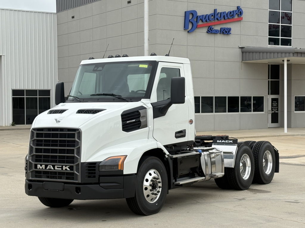 Mack AN64T conventional daycab truck in white, shown in dealership lot with Brunkner's building in background, front grille with MACK badge, dual rear axles.