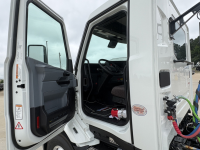 Truck cab interior view with open door of a white Mack AN64T. Steering wheel visible, dashboard, and seat. Exterior shows door label and small circular badge.