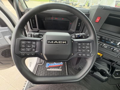 Mack truck steering wheel with controls and dashboard visible; center shows 'MAC K' logo, floor mat reads 'Bruchneck' or dealership sticker; surrounding controls and dashboard buttons visible.