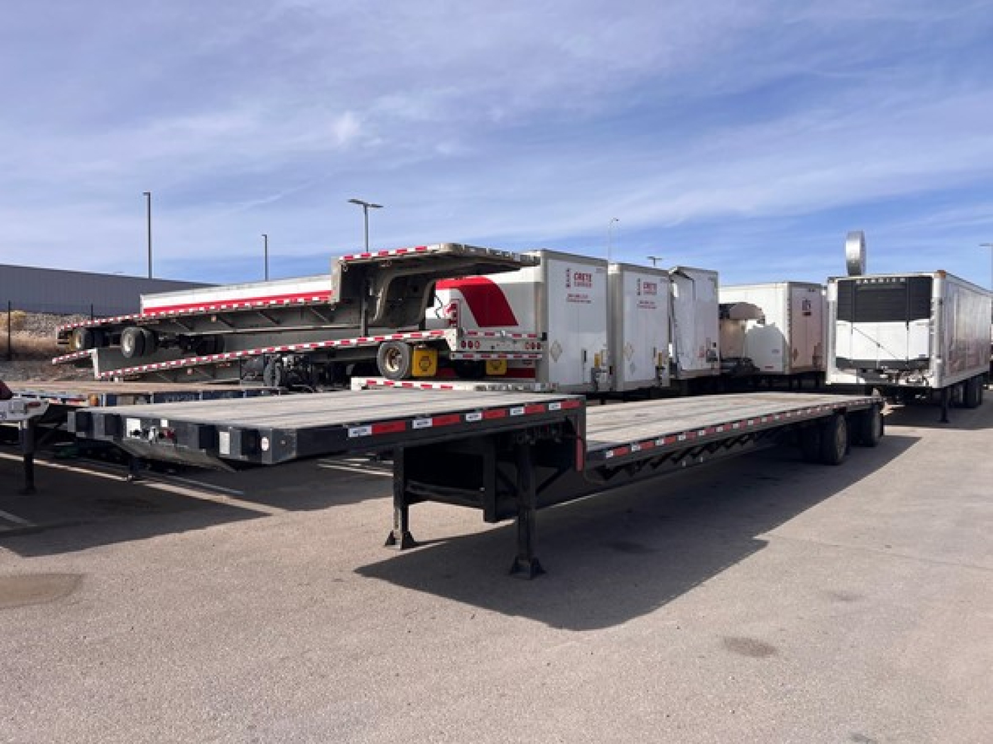 Trailer deck showing long flatbed with loading ramp and three enclosed trailers in background; visible branding on equipment.