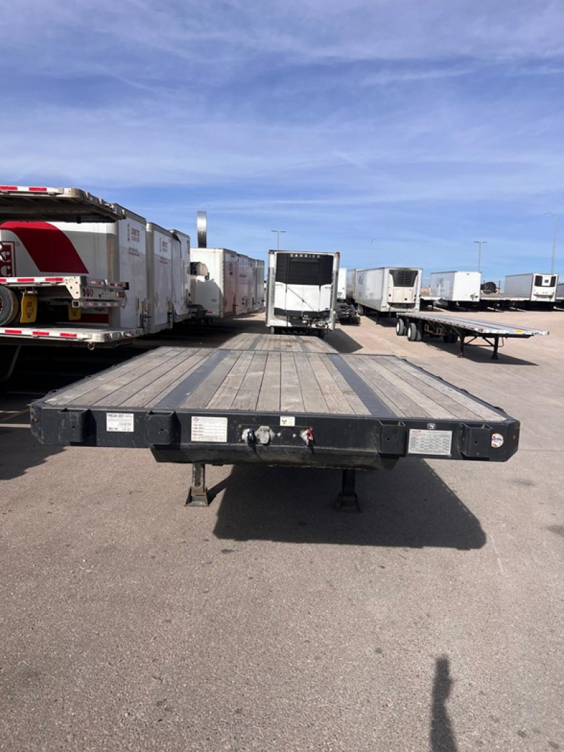 Flatbed trailer with wooden deck and metal frame, front coupler visible; multiple trailers parked in background; clear sky.