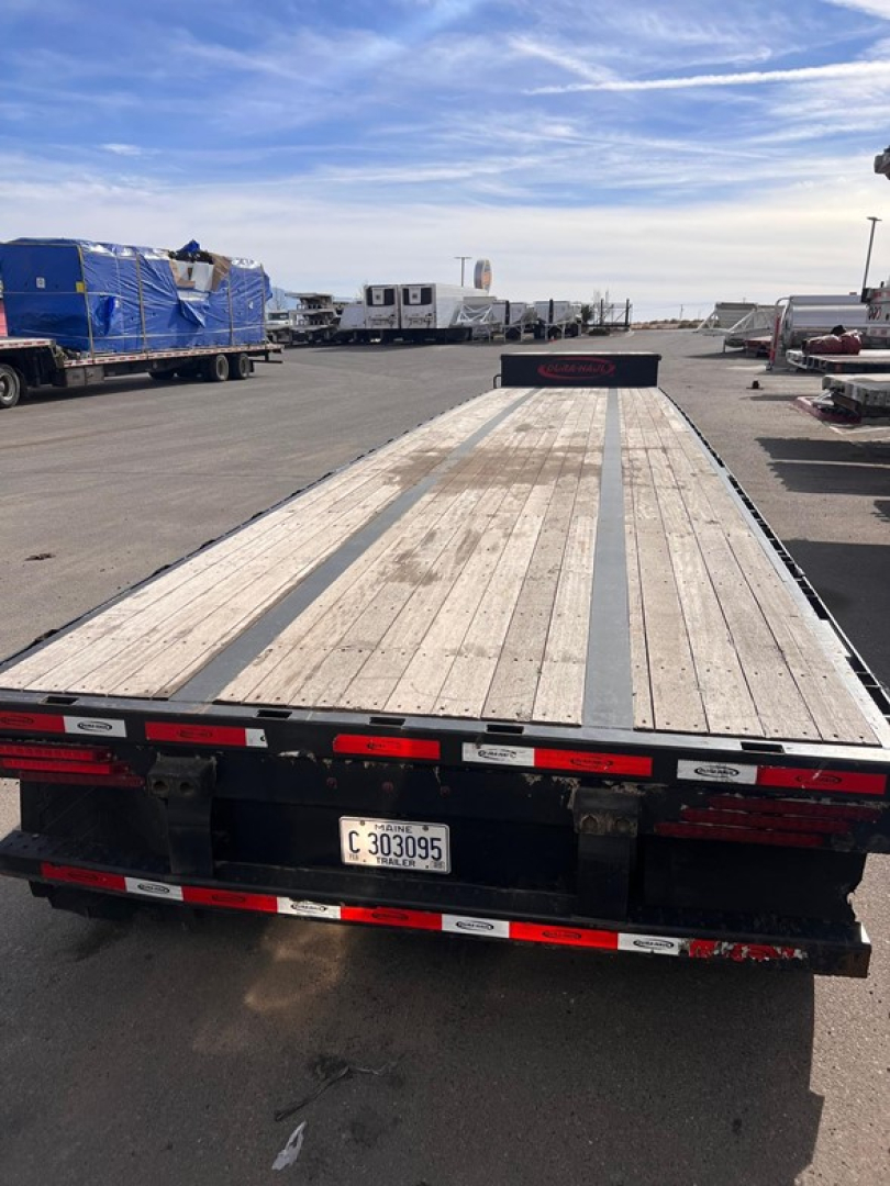 Trailer rear view showcasing a long drop deck trailer with wooden planks, black frame, red-and-white safety markings, license plate C303095, and fleet yard background.
