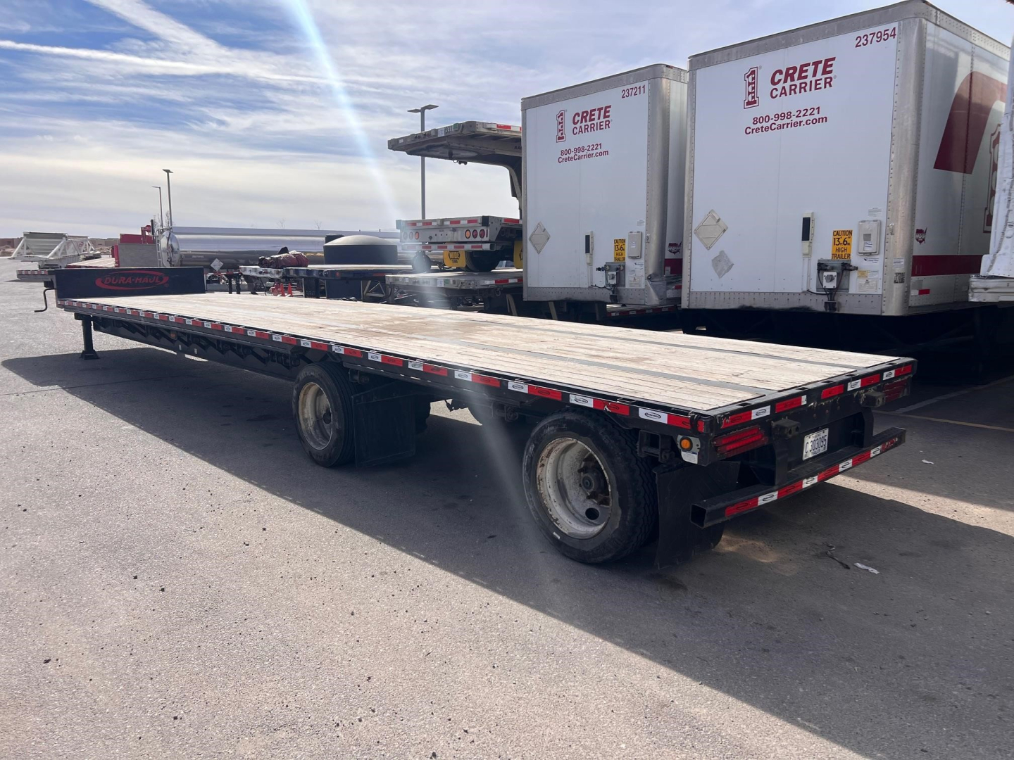 Flatbed trailer showing long wooden deck, tandem axle, and rear lights; two rear doors of nearby trailers in background with Crete Carrier logos.