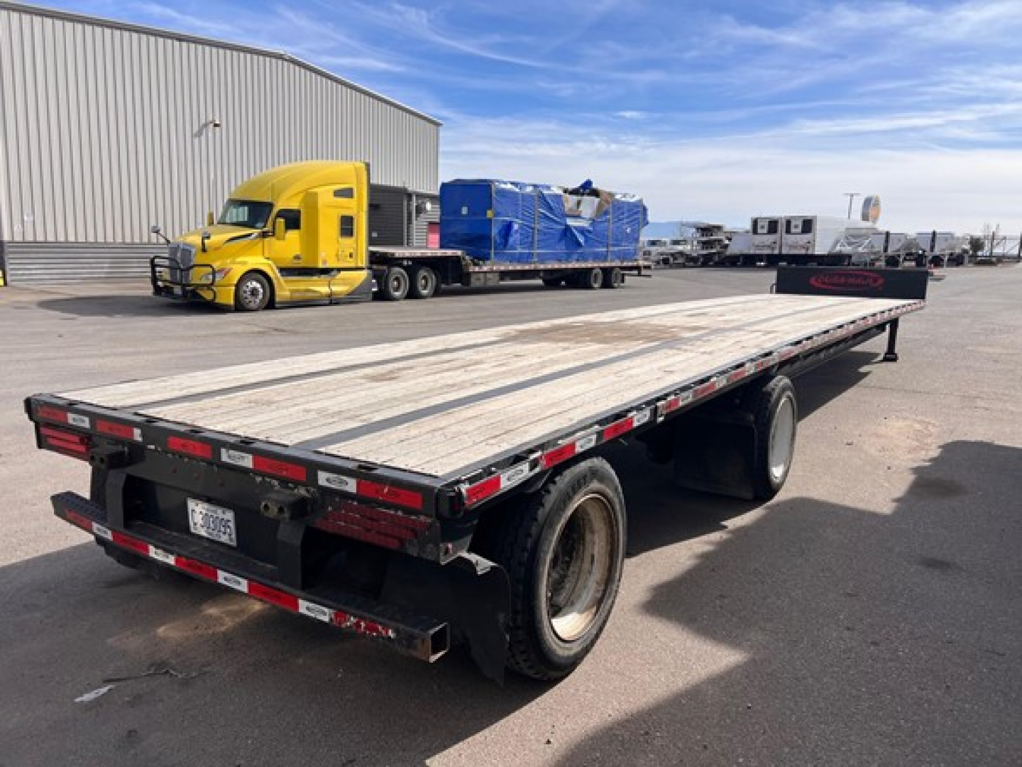 Flatbed trailer with wooden deck and dual rear axles, parked in lot beside a yellow truck and other trailers; long chrome rails with reflective tape.