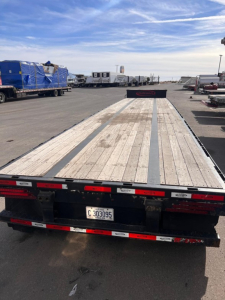 Trailer rear view showcasing a long drop deck trailer with wooden planks, black frame, red-and-white safety markings, license plate C303095, and fleet yard background.