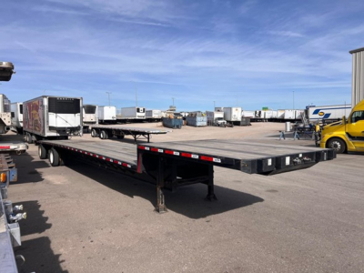 Flatbed trailer in a yard showing a drop-deck configuration with low-profile frame and a gooseneck-style front. Visible text: none legible on the trailer itself.