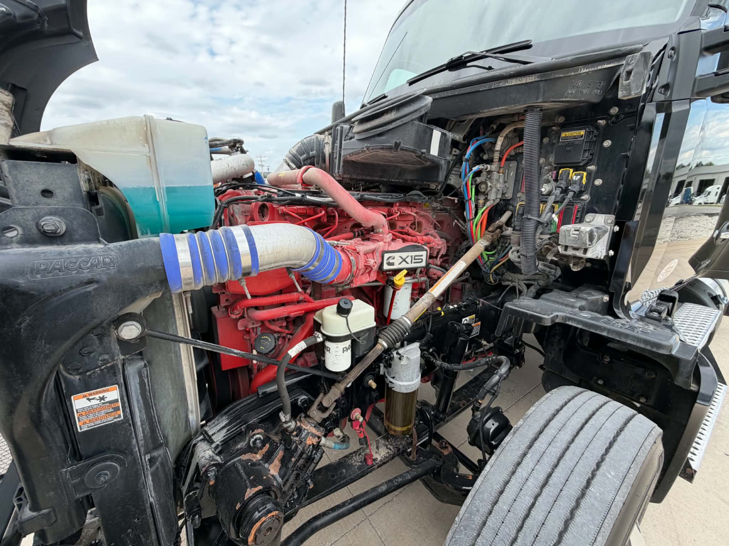Kenworth T680 engine bay showing a red Cummins ISX engine with blue silicone coupler, wiring harness, coolant reservoir, and exposed components.