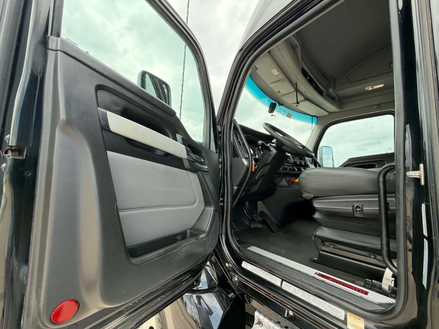 Kenworth T680 cab interior showing driver seat and steering wheel with open door; dashboard and pedals visible.