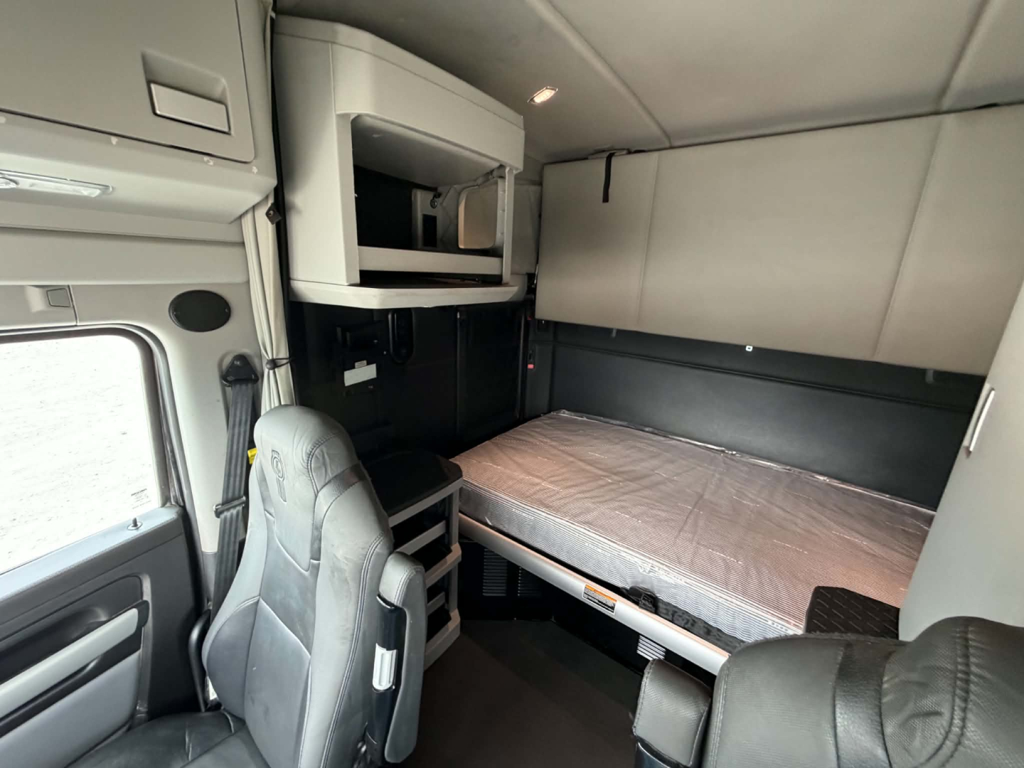 Kenworth T680 sleeper cab interior showing bunk bed, driver seat, overhead storage, and window; compact cabin with light grey panels.