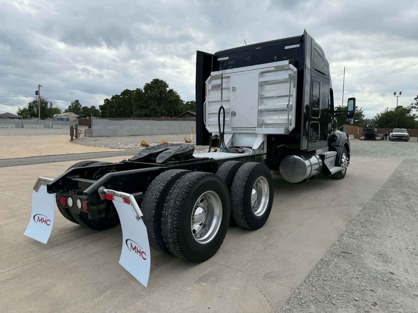 Kenworth T680 tractor with rear view showing fifth wheel, dual rear axle, and side fuel tanks. Visible text on white mud flaps reads MHC.
