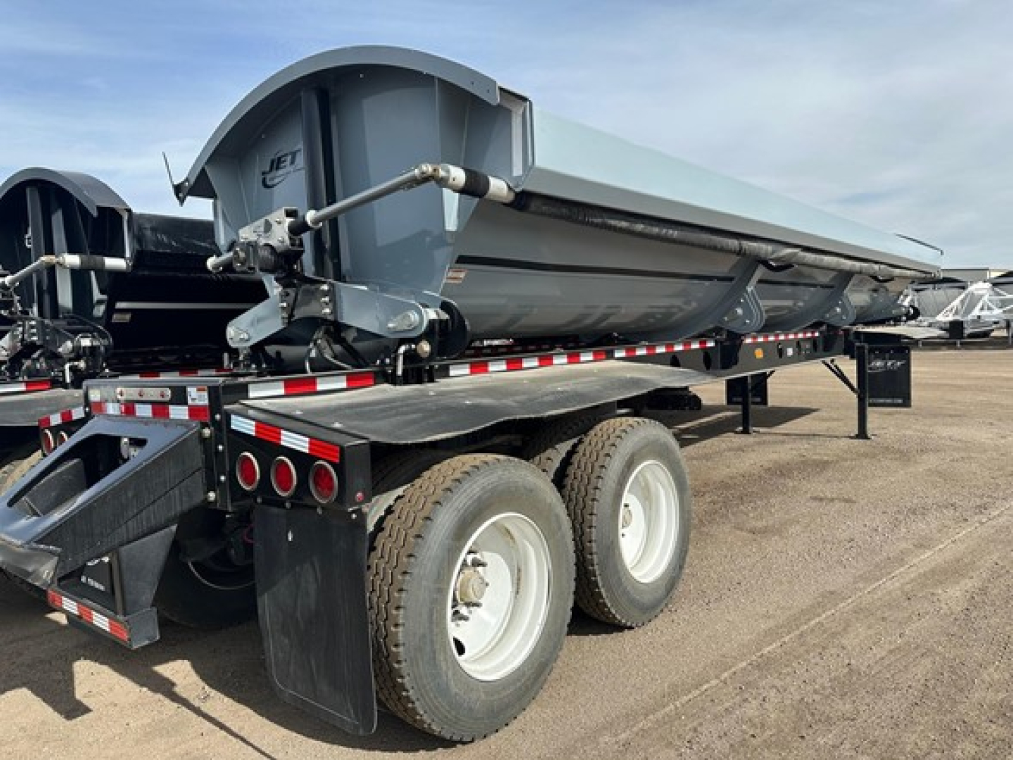 Jet Air Ride side dump trailer showing rear discharge chute and hydraulic piping, dual rear axles with large tires, gray dump body and black undercarriage.