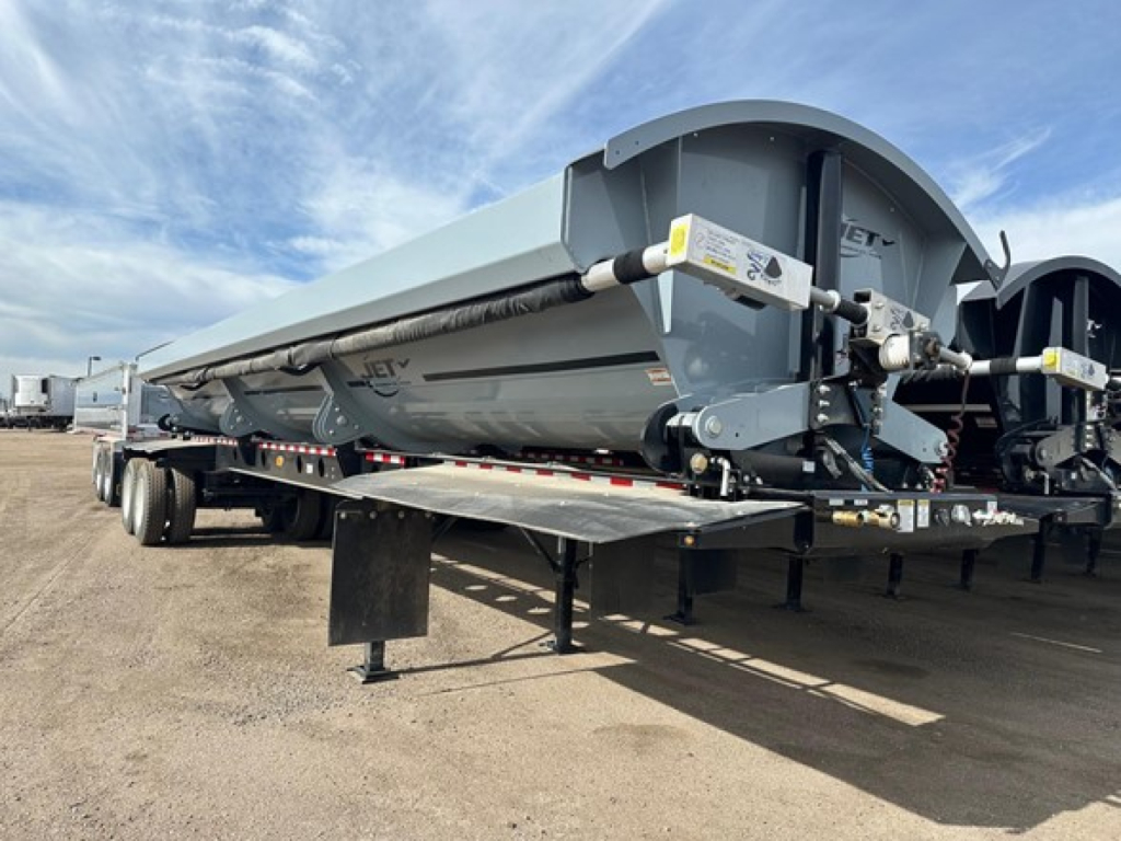Jet Air Ride Side Dump trailer in a yard, side-view showing the long grey body, support legs, and dual rear wheels with hydraulic components and safety decals visible.