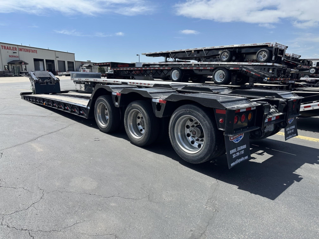 Load King RGn lowboy trailer with triple-axle rear, flatbed sections, and ramp/auger visible; other trailers stacked in background.