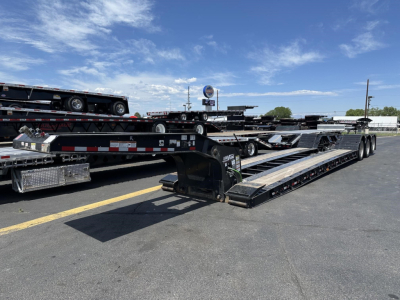 Lowboy trailer with long loading deck and ramps, multiple axles, Load King branding visible on frame; clear sky, other trailers in the background.