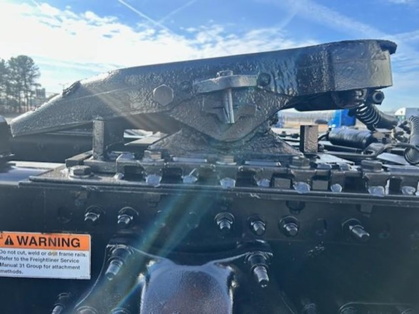 Tractor-trailer truck hitch/frame with hydraulic fifth-wheel and mounting bolts on Freightliner Cascadia, left front view showing mounting assembly and warning label.