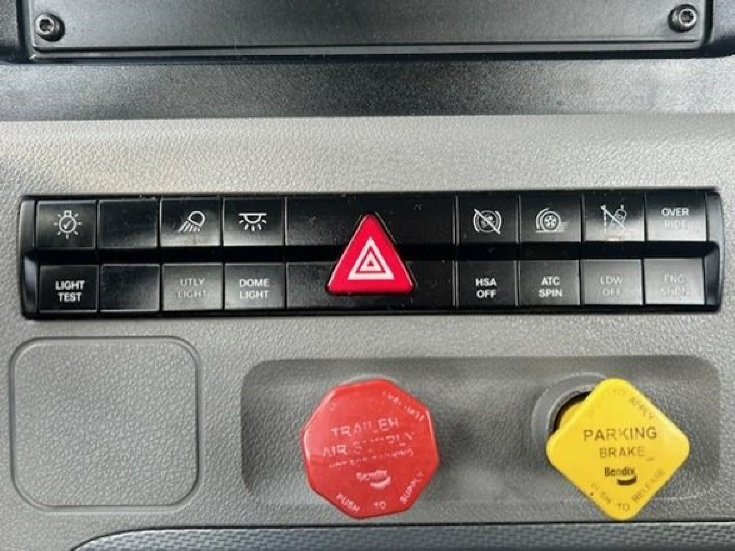 Freightliner Cascadia truck interior panel showing hazard warning triangle button, light controls, and red trailer air line knob with yellow parking brake knob nearby.