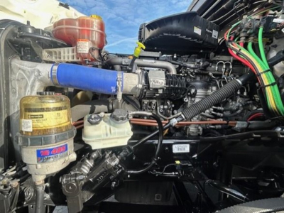 Diesel truck engine bay with blue coolant hose, yellow reservoir, and surrounding components in a Freightliner Cascadia engine compartment.
