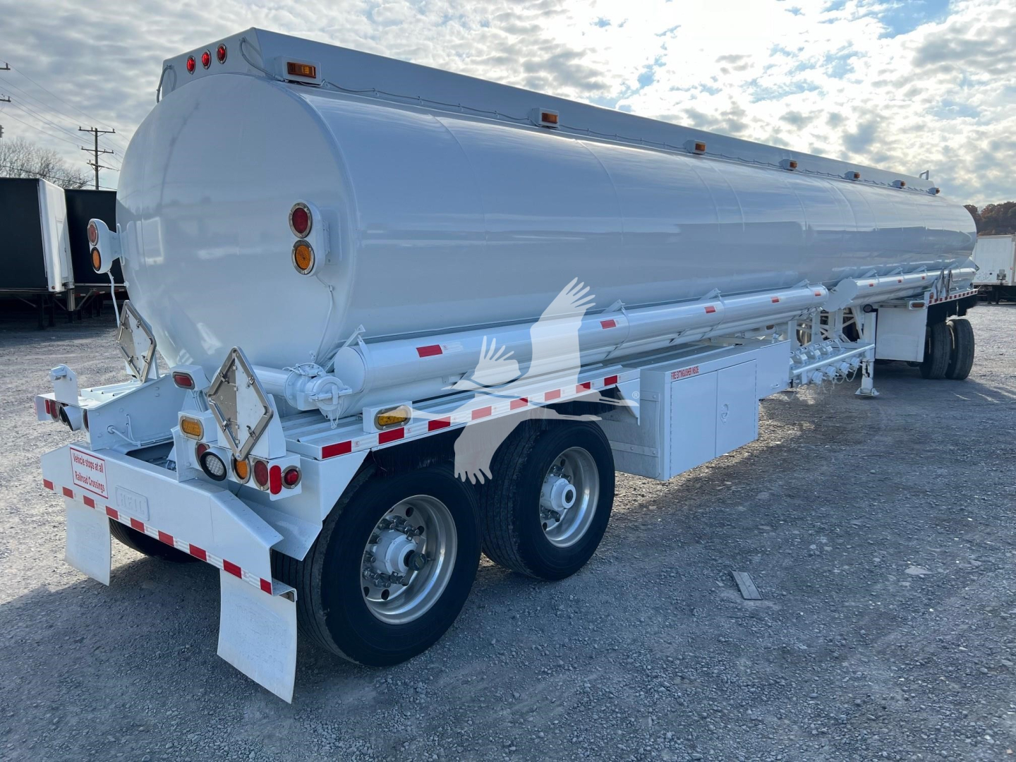 Petro tank trailer showing cylindrical white Heil trough bottom fuel tank with dual rear axle, rear lights, and support ladder; side access compartments; VIN tag not visible.