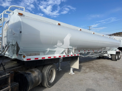2013 Heil trough bottom petro tank trailer viewed from side, white cylindrical tank with dual rear axles and support legs, black fuel tank front, safety rails and reflective tape along sides.