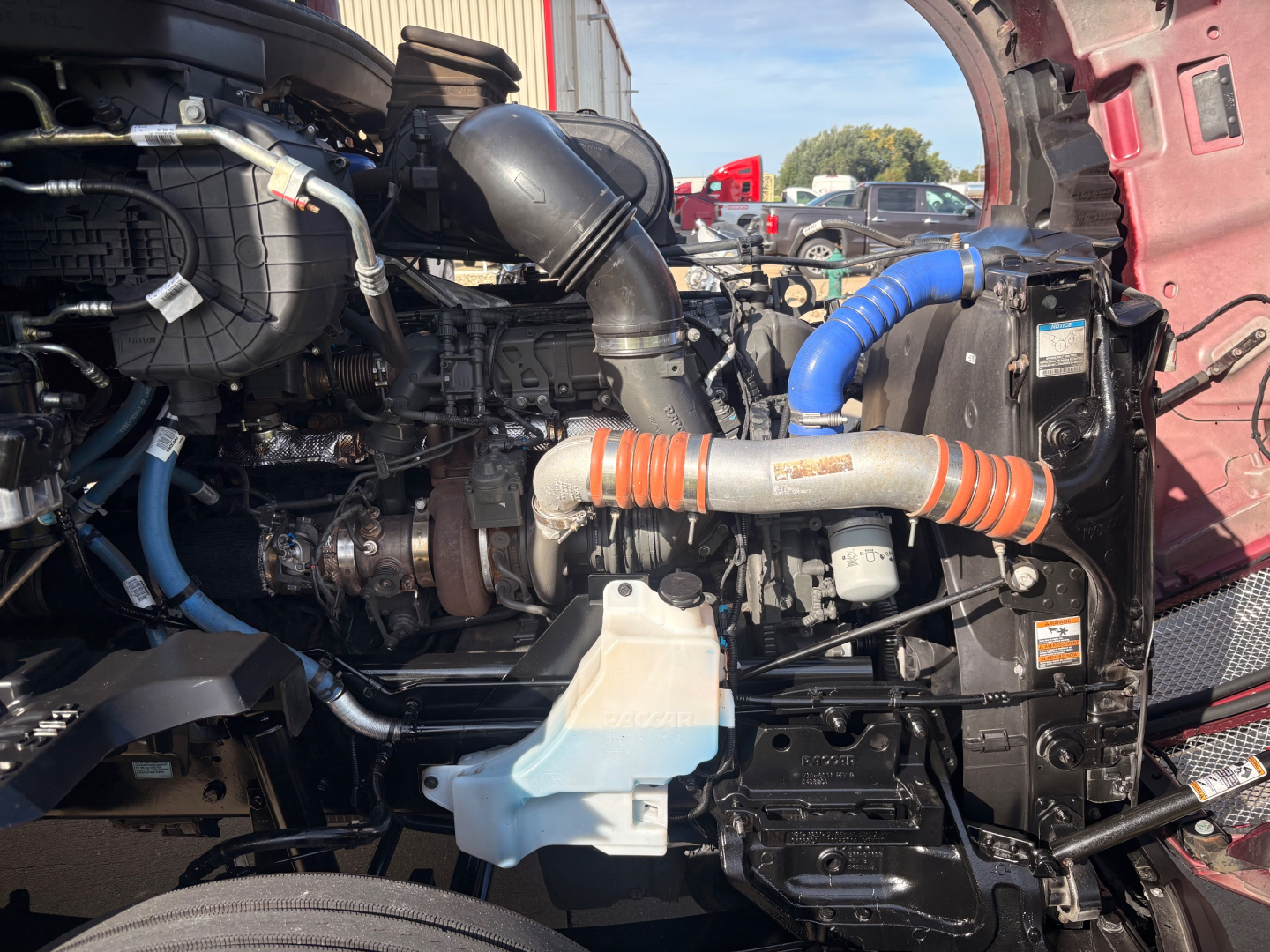 Kenworth T680 engine bay showing blue hoses, orange-striped intercooler hose, coolant reservoir, and various engine components.