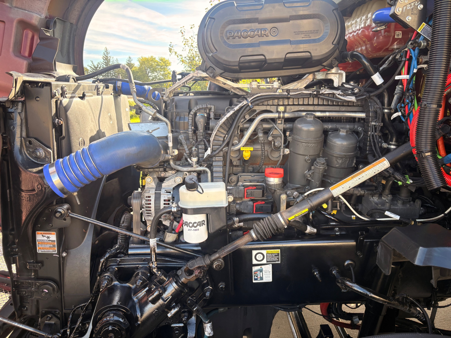 Kenworth T680 engine bay showing PACCAR MX-13 engine with intake tubes, coolant reservoir, and various hoses and wiring in view.
