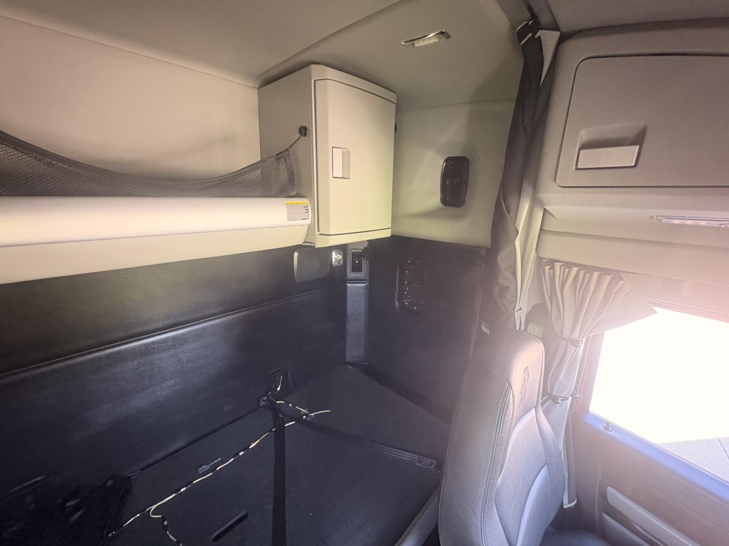 Kenworth T680 sleeper cab interior showing upper bunk and storage cabinet with electrical panel, window with curtain, and seat in cabin.