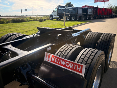 Kenworth truck chassis in foreground with Kenworth badge and dual rear axles; yard and trucks in background.