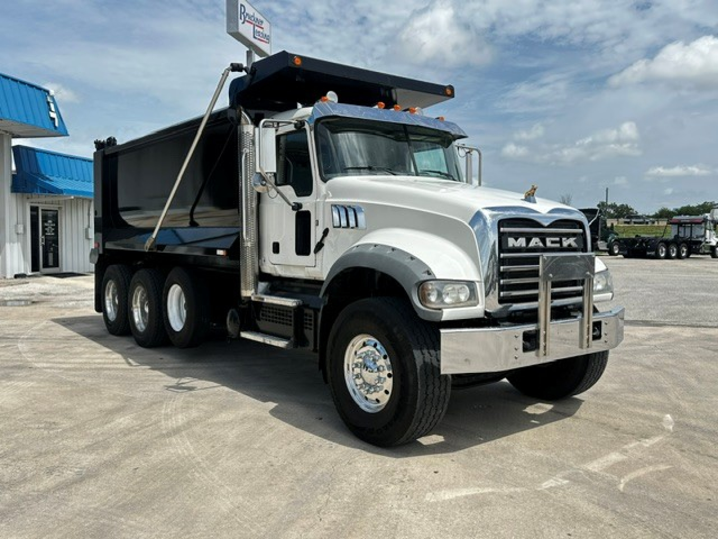 Mack GR64F dump truck with silver cab and black dump body, front grille with MA C K letters, dual rear axles, chrome bumper, on concrete lot.