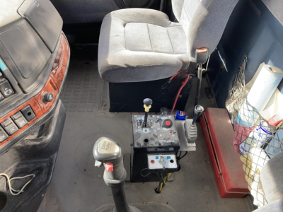 Volvo truck cabin interior with gear selector lever, joystick controls, and a seat. Center console shows gear shifter and control panel; pedals and dashboard partially visible.