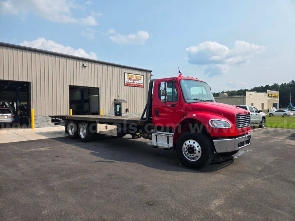 Red Freightliner M2 tow truck in front of a service shop with flatbed showing seating cabin and wheels.