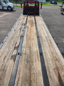 Flatbed tow truck bed with wooden planks and metal rails, rear cab in background, Freightliner M2 listing details visible in context.
