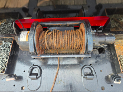 Truck winch with metal spool and wound wire rope mounted on a flatbed, showing hooks and mounting plate, in-service condition.