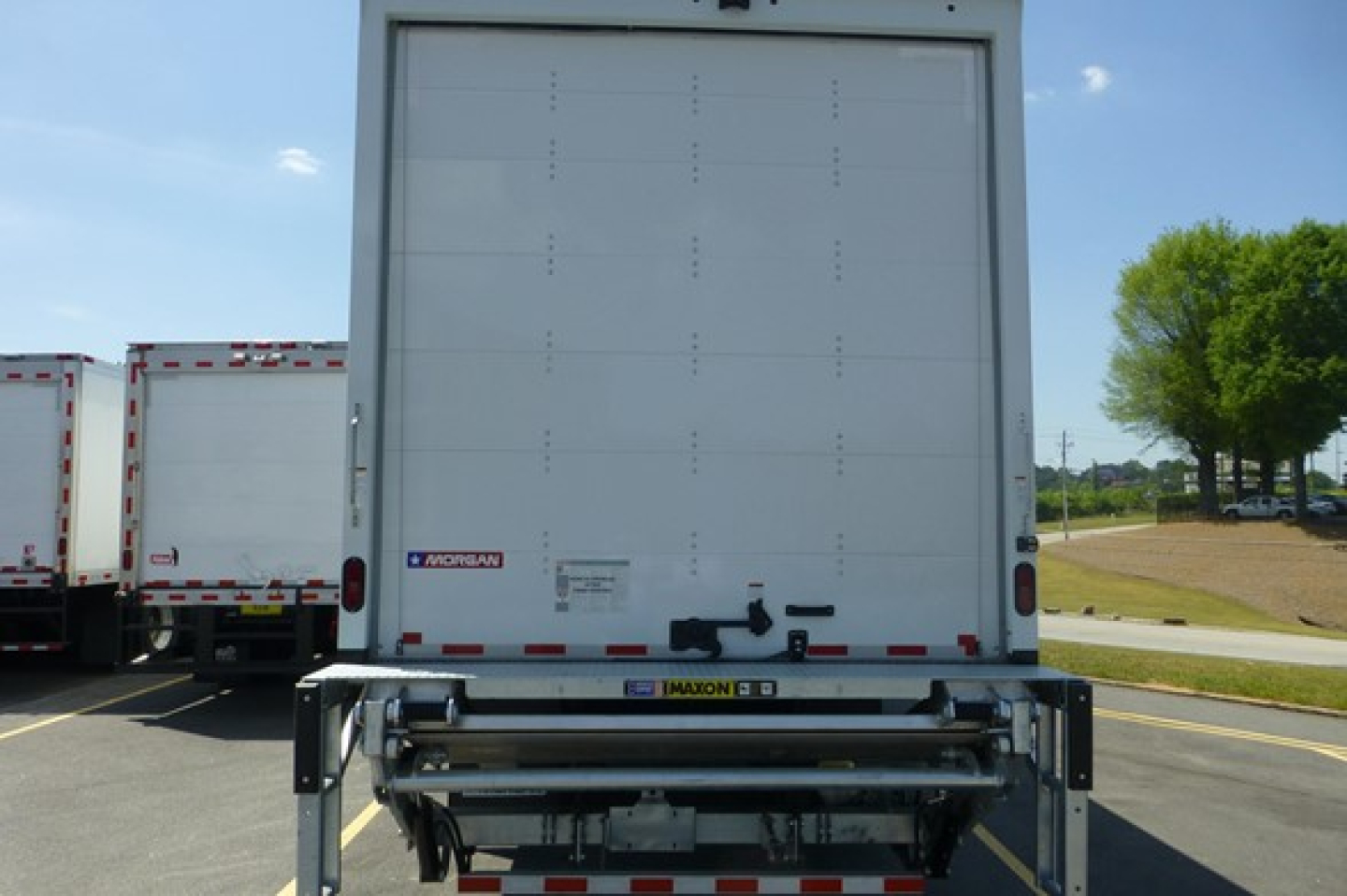 Rear view of a Freightliner M2 106 box truck with a roll-up rear door and hydraulic lift, visible VIN/stock stickers; clear sky and parking lot in background.