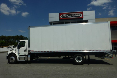 Freightliner M2 106 box truck in-side view showing cab and large box with Freightliner dealership building in background.