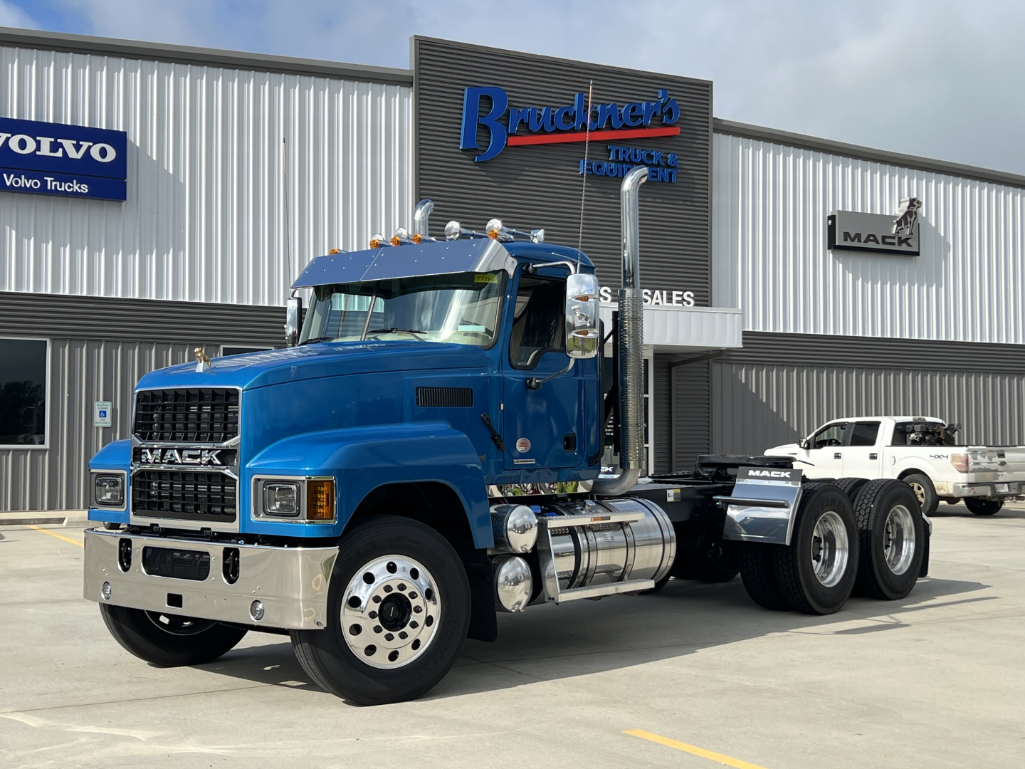 Blue Mack PI64T tractor with chrome grille and dual rear axles, parked in front of a dealership with Mack and Volvo signs; visible text includes MACK on grille and dealership signs.
