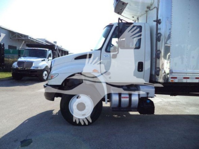 Truck engine bay view of a white 2019 International Durastar 4300 refrigerated truck cab with large side mirror and fuel tanks visible; background shows another vehicle.