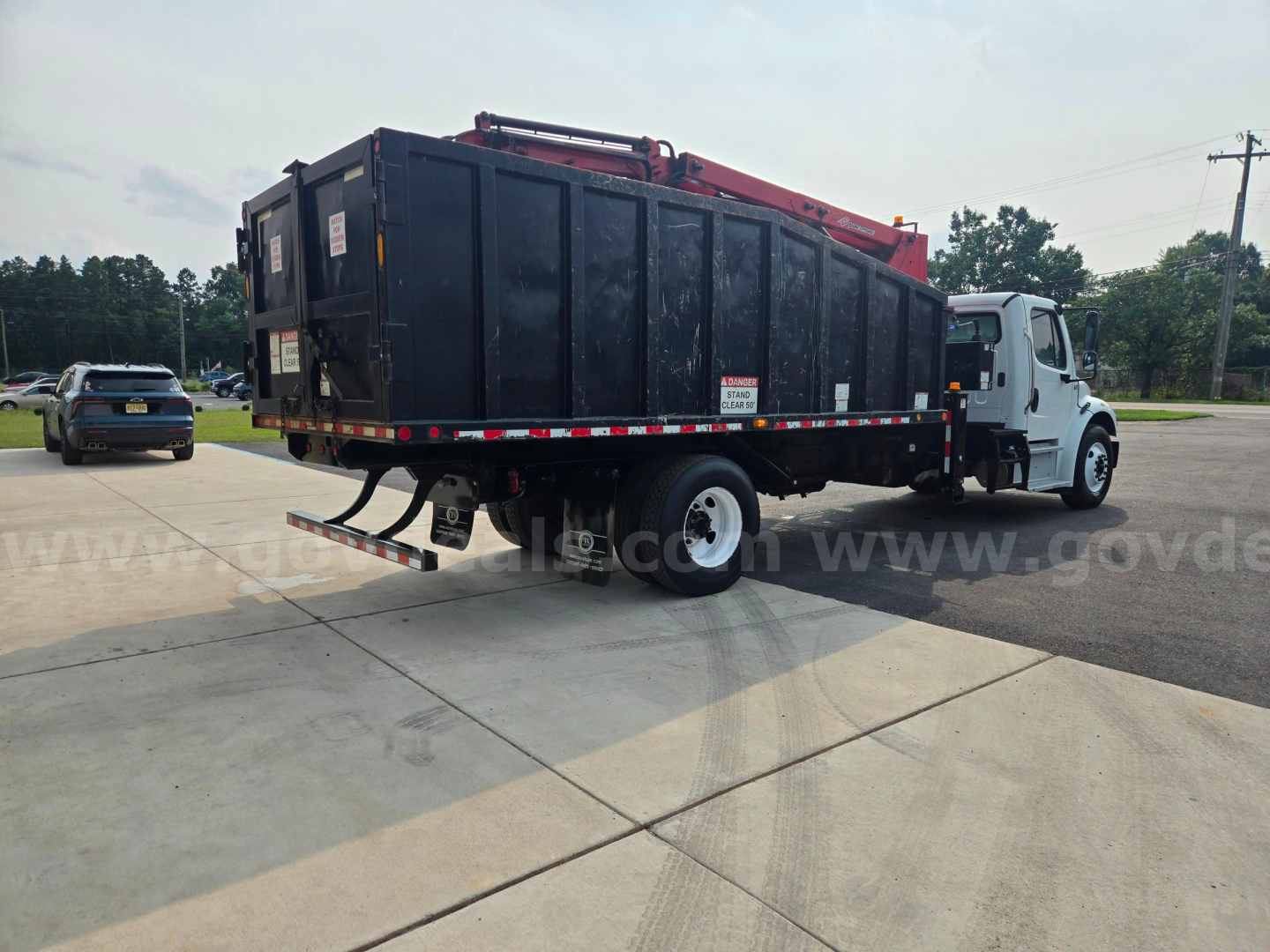 Truck with large black grapple dumpster body mounted on a white Freightliner M2 cab showing side view of cargo area and rear bumper.