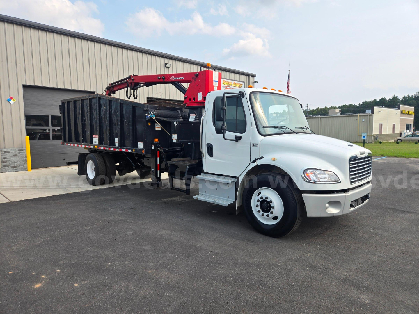 Truck side view of a 2019 Freightliner M2 grapple truck with a red crane and black hopper/storage bed, white cab, in a dealership lot.