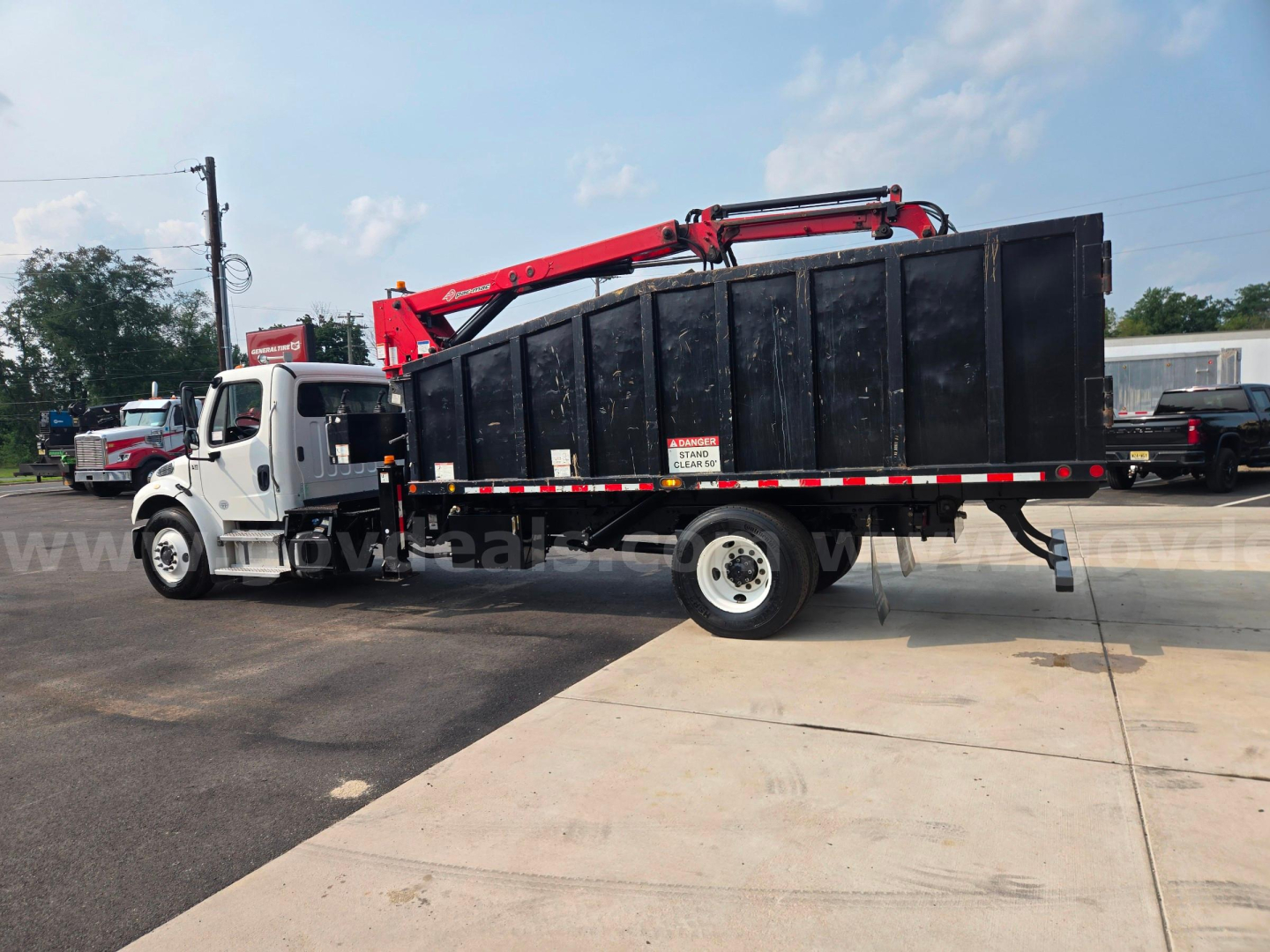Truck equipped with a black dump body and red hydraulic crane/overhead grapple on a Freightliner M2 chassis, side view showing stock number 29174-85 and multiple warning decals.