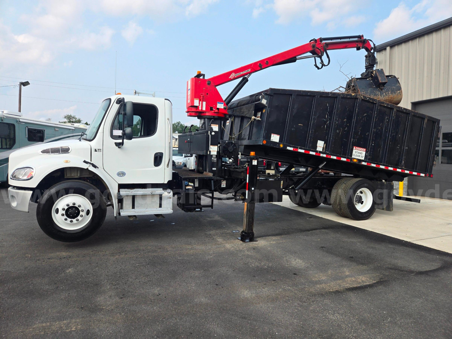 2019 Freightliner M2 grapple truck showing crane with red articulated arm loading a large black dumpster into a dump bed.