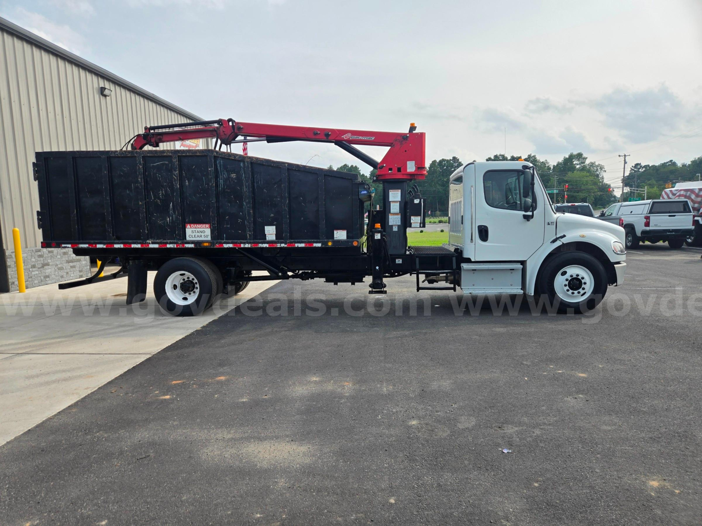Truck side view showing a white Freightliner M2 with a black dump body and red hydraulic crane arm for grappling loads.