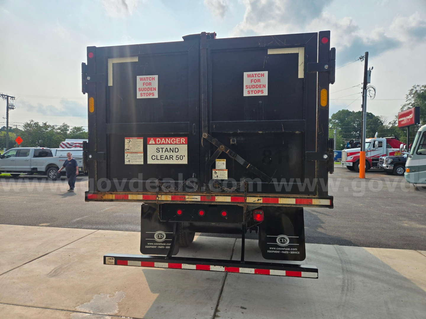Freightliner M2 dump truck rear showing safety decals and bumper; two rear doors with signs: 'DANGER STAND CLEAR 50''; additional labels 'WATCH FOR SUDDEN STOPS' on each door; rear underride guard and tail lights visible.