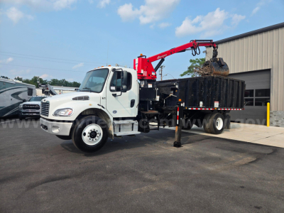 2019 Freightliner M2 grapple truck with crane arm and long black dump bed, front cab white, parked outside a building