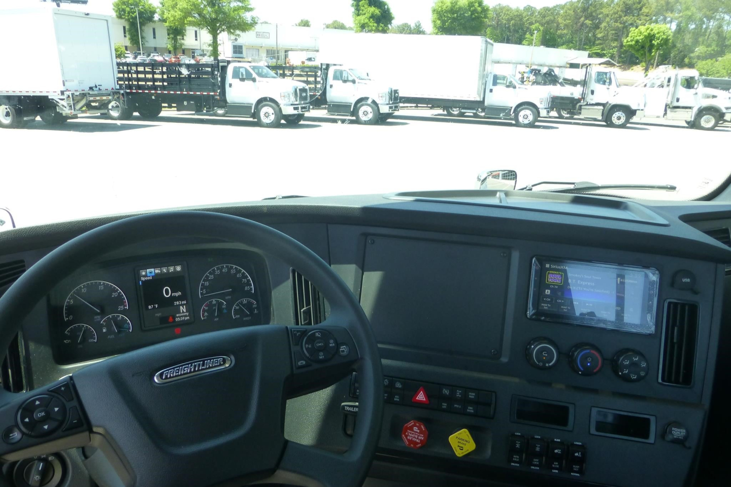 Freightliner Cascadia truck interior dashboard with steering wheel, instrument cluster, and navigation display, view through the windshield of a truck lot with multiple trailers.
