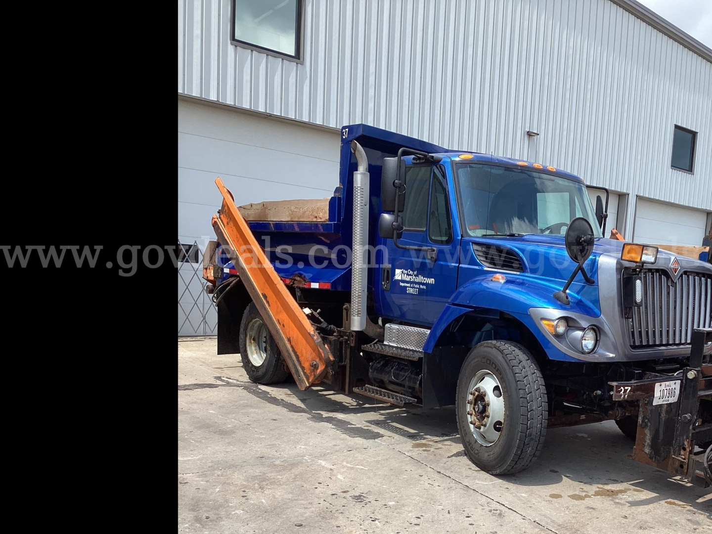 2016 International 7300 plow truck in blue with orange plow and hydraulic bed, front-side view showing side mirrors and wheel fenders.
