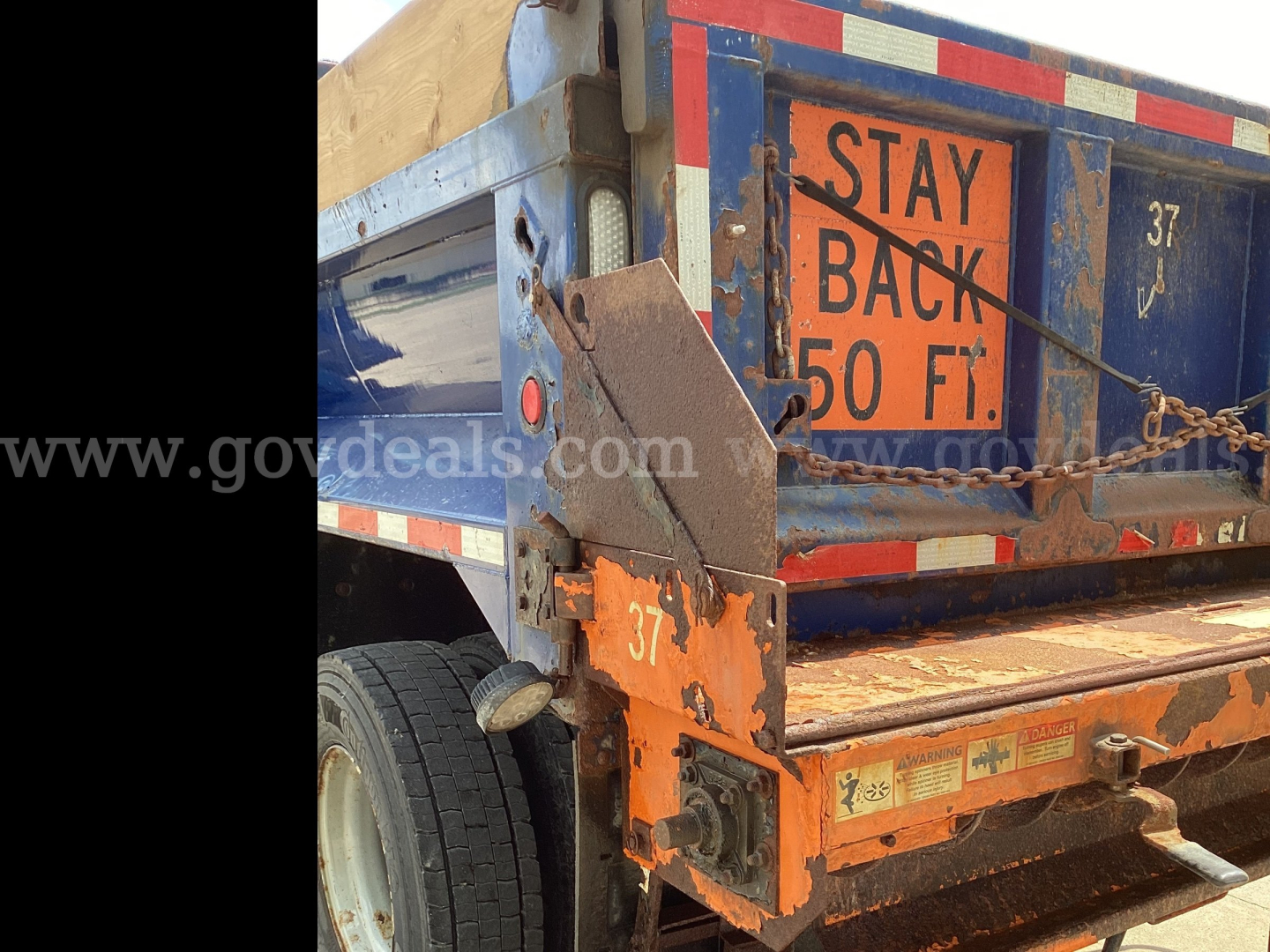 Rear view of a blue plow truck showing safety sticker and orange rear bumper with chain and stay back 50 ft sign.