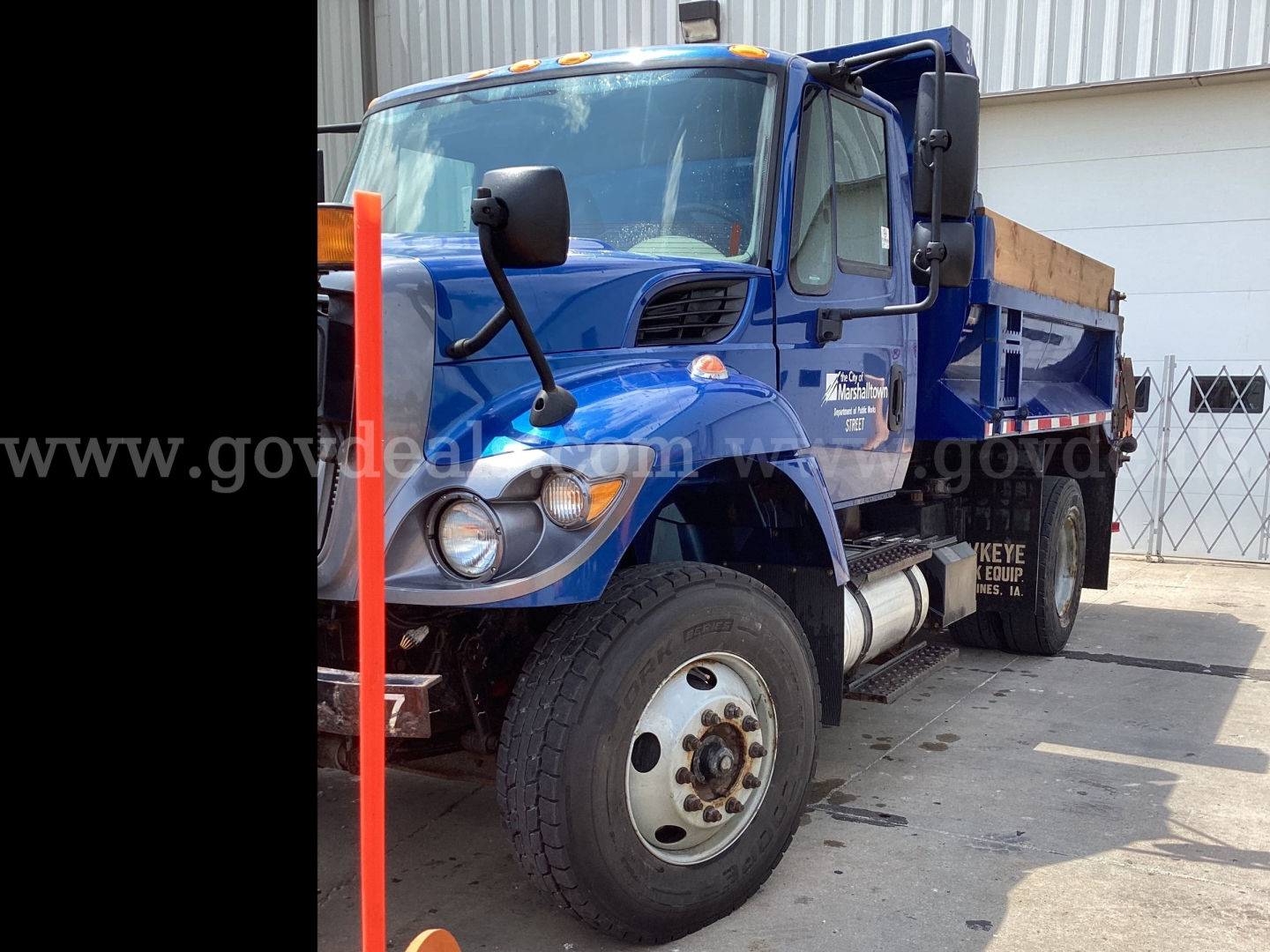 Truck cab in view showing International 7300 plow truck with blue color, front wheel, side mirrors, and dump bed partially visible.