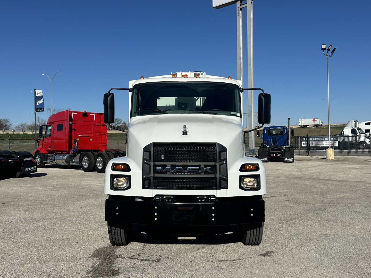 Front view of a white 2026 Mack MD6 truck in a dealership lot, showing grille with Mack emblem, large bumper, and side mirrors.