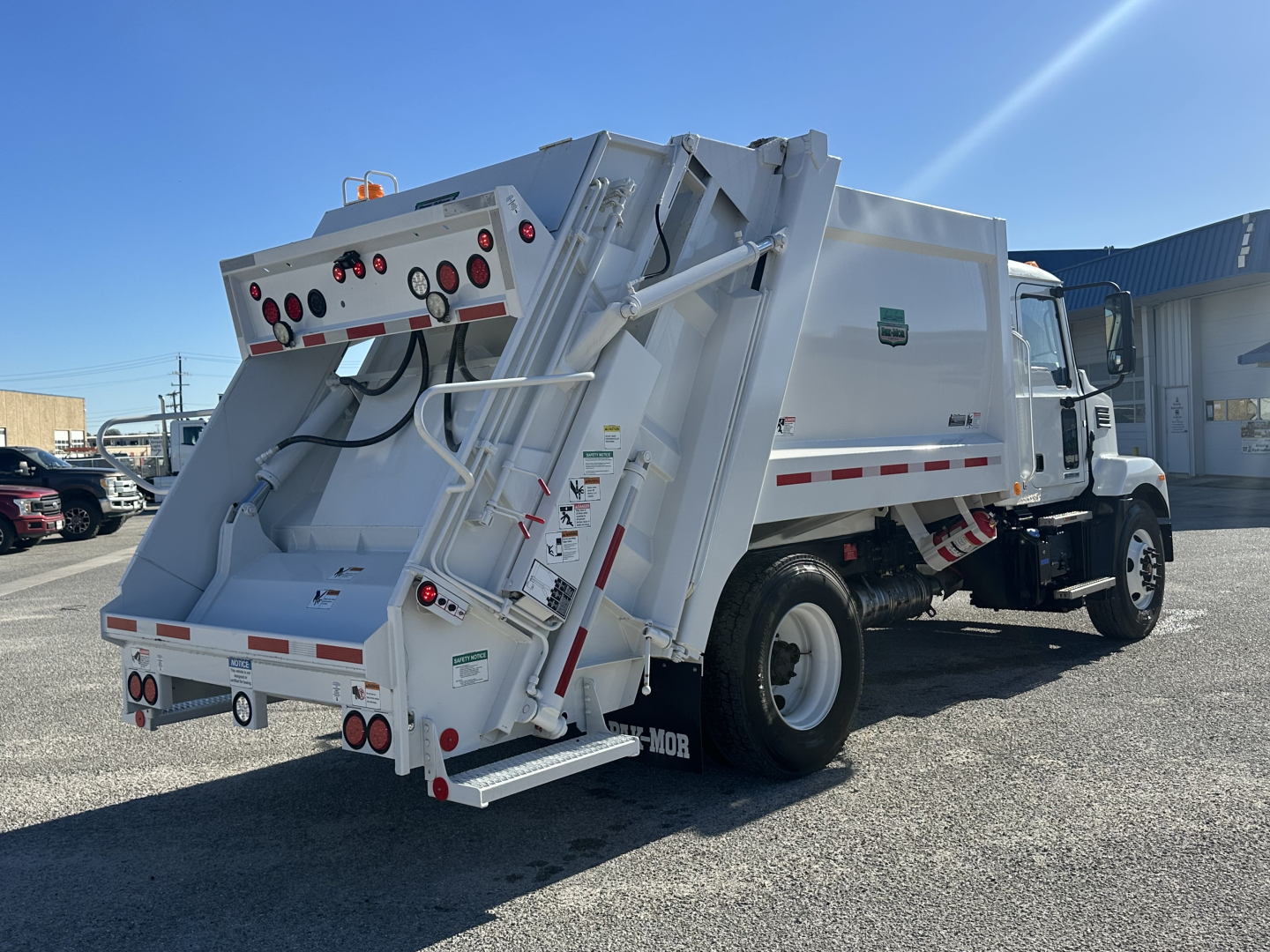 Mack MD6 rear unload refuse truck in service-ready condition, white body, rear hopper with hydraulic arms and controls, side cab, visible decals and warning labels.